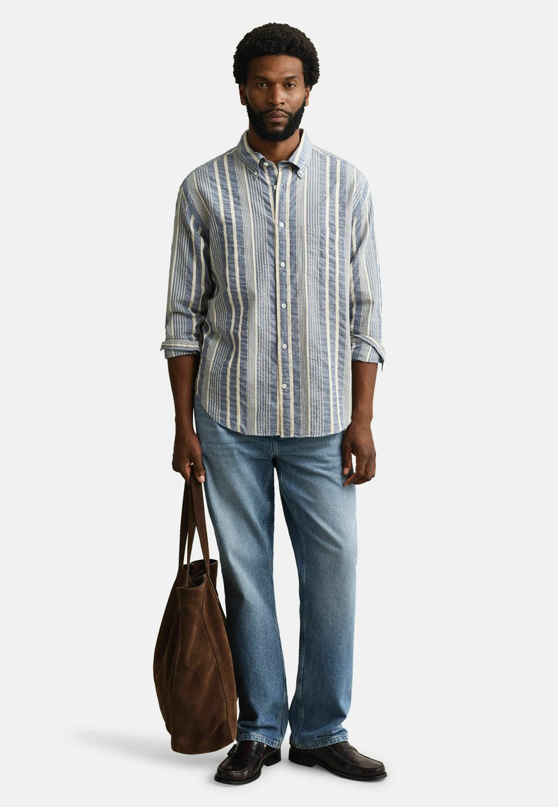 Mann trägt blau-weiß gestreiftes Hemd, hellblaue Jeans, schwarze Schuhe und hält eine große braune Wildleder-Shoppertasche an seiner Seite.