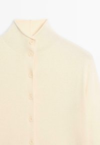 Ljus krämfärgad cardigan med hög krage, prydd med sex matchande knappar framtill. Mjuk textur med ett lätt ribbat stickmönster.