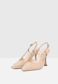Beżowe, lakierowane szpilki typu slingback z ostrym noskiem, smukłym paskiem na kostkę oraz obcasem w szpilkę. Gładka faktura i prosty design.