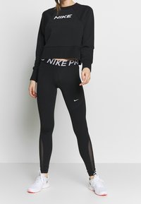 Sudadera corta negra con el logo blanco de "NIKE", combinada con leggings negros que tienen una cinturilla "NIKE PRO" y cortes de malla. Zapatillas blancas.