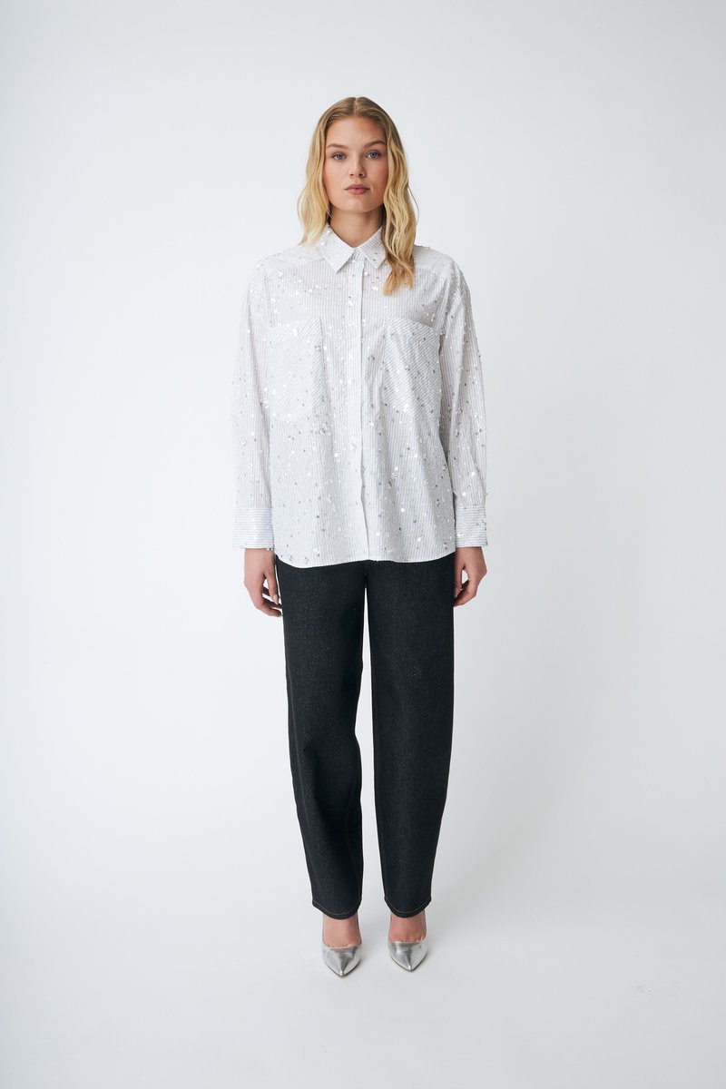 Camicia bianca con un delicato motivo a righe e dettagli riflettenti, abbinata a pantaloni scuri a gamba larga e scarpe argentate con punta.