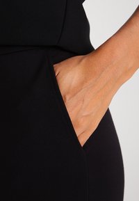 Vêtement noir à coupe ajustée avec une poche latérale. Une main est glissée dans la poche, mettant en valeur la texture lisse du tissu.