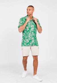 Groen shirt met korte mouwen met witte tropische bladeren, gecombineerd met beige shorts en witte sneakers. Eenvoudig ontwerp met een relaxte pasvorm.