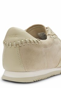 Baskets en daim et cuir beige avec des détails cousus au talon et une semelle blanche texturée, présentées en angle pour montrer l'arrière et le côté.