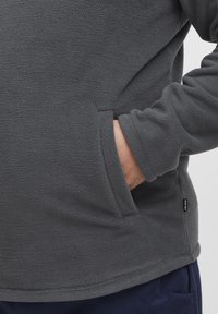 Grauer Fleece-Pullover mit weicher, strukturierten Oberfläche, ausgestattet mit einer Kängurutasche und einem kleinen Logo-Tag an der Seite, entworfen für Komfort.