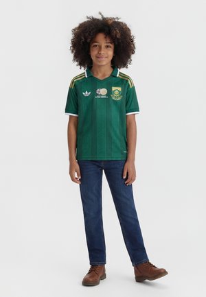 Copil cu păr creț purtând un tricou verde de fotbal al Africii de Sud, blugi albaștri și cizme maro, stând pe un fundal simplu.