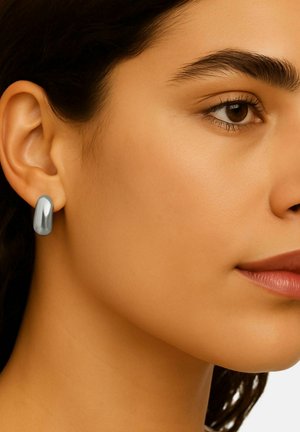 Boucle d'oreille en métal argenté présentant une forme lisse et arrondie avec une finition polie, portée à l'oreille d'une personne.