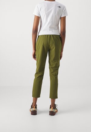 Person trägt olivgrüne kurze Hose, weißes kurzärmeliges T-Shirt mit schwarzem Logo und Wanderschuhe, steht mit dem Rücken zur Kamera vor weißem Hintergrund.