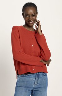 Lächelnde Frau mit geflochtenem Haar trägt einen roten Knopf-Pullover und blaue Jeans, posiert mit einer Hand, die ihr Gesicht berührt.