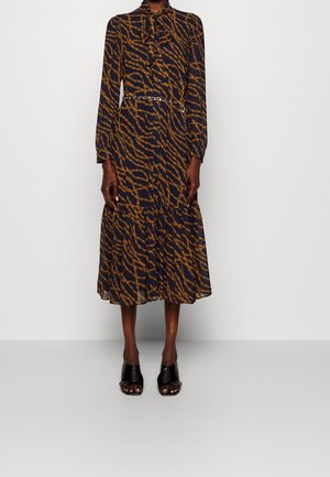 Femme portant une robe midi noire à manches longues avec des motifs de chaînes marron, un col noué et des chaussures à talons ouvertes noires.
