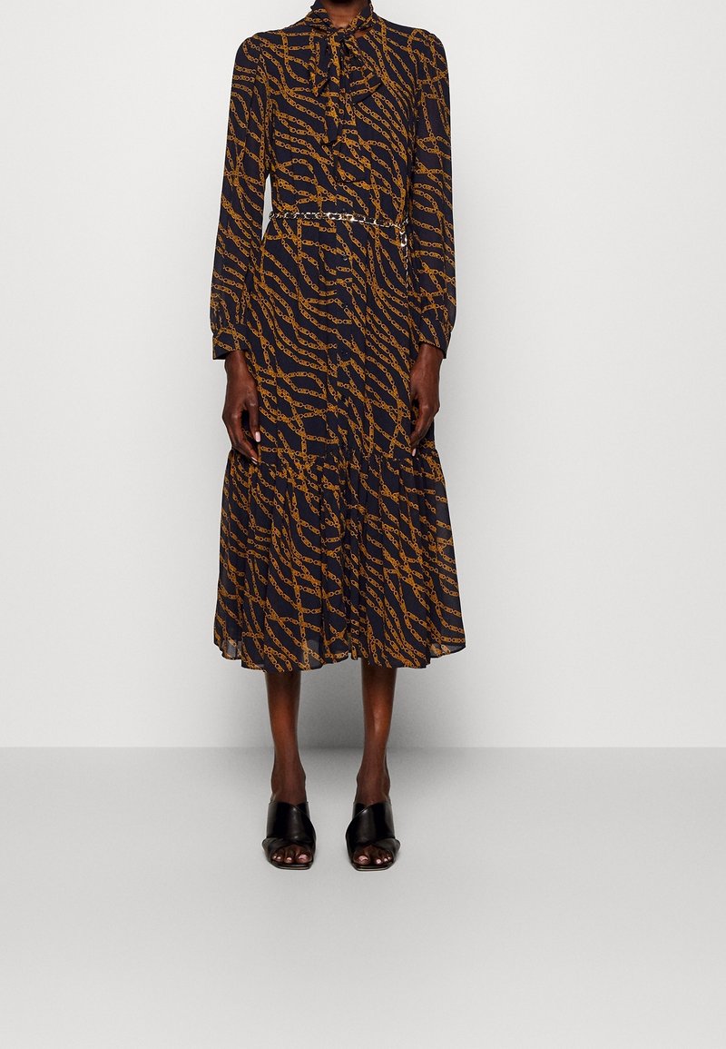 Femme portant une robe midi noire à manches longues avec des motifs de chaînes marron, un col noué et des chaussures à talons ouvertes noires.