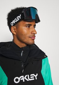 Skibril met een zwarte band met een wit "OAKLEY" logo, blauwe getinte lens, gedragen door een persoon met een zwart en groene jas.