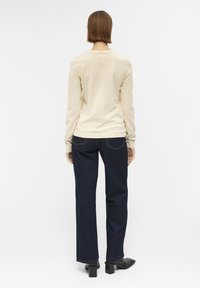 Un haut beige à manches longues et un jean large bleu foncé. Le haut a une texture lisse, tandis que le jean présente des coutures contrastantes et des poches.