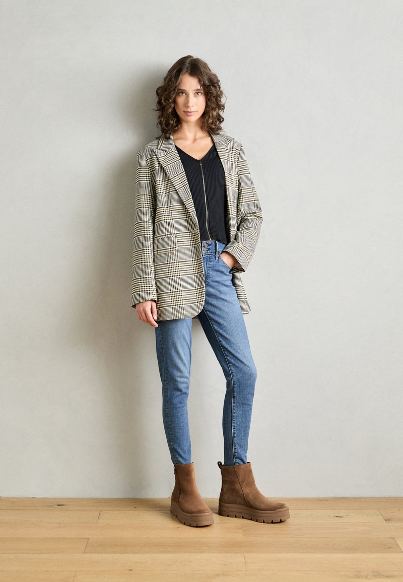 Blazer de cuadros en beige y negro, combinado con una camiseta de canalé negra, jeans ajustados azules y botines de ante marrón con suelas gruesas.