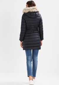 Black puffer coat with a fur-lined hood, ribbed texture, fitted waist, and front zip closure, paired with blue jeans and white shoes.