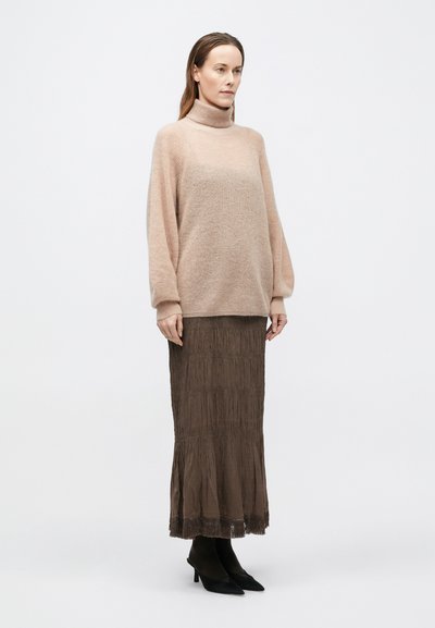 Puha, halványpink, túlméretezett kötött pulóver magas nyakkal; barna, hajtott, földig érő szoknya rojtos szegéllyel; fekete, hegyes orrú magassarkú cipő.