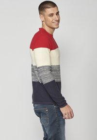 Maglione lavorato a maglia con righe orizzontali rosse, crema, grigie e blu navy; texture a coste, collo a giro, abbinato a jeans blu.