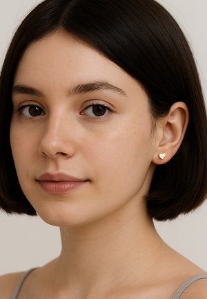 Giovane donna con capelli corti e scuri che indossa piccoli orecchini a forma di cuore dorati, che guarda leggermente di lato su uno sfondo neutro.