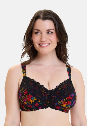 Mujer sonriente que lleva un sujetador de encaje floral negro con patrones de flores coloridas y tirantes ajustables, mirando ligeramente hacia un lado.
