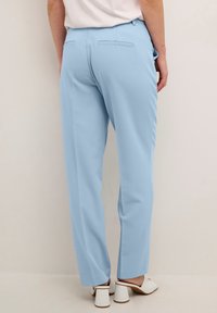 Pantaloni tailored blu chiaro con una texture liscia, dotati di tasche posteriori e gamba leggermente affusolata, abbinati a mule con tacco bianchi.