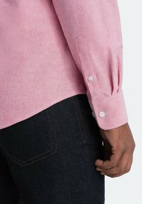 Vaaleanpunainen pellavapaita, jossa on rento leikkaus, valkoiset napit ja teksturoitu kudos, yhdistettynä tummiin farkkuihin.