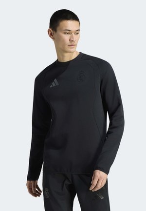 Mann trägt schwarzes langärmliges Sportoberteil und passende Hosen mit dunklen Logos, schaut nach links vor einem schlichten Hintergrund.