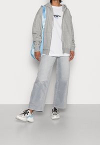 Sweat à capuche gris clair zippé avec cordon de serrage, porté par-dessus un T-shirt blanc. Associé à un jean bleu clair à jambes larges et des baskets multicolores.