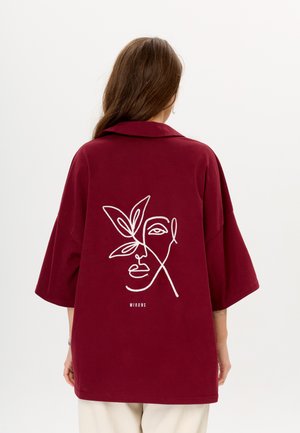 Bordeaux oversized shirt met een geknolde kraag. Heeft witte geborduurde lijntekening van een gezicht en een blad op de achterkant. Tekst "MIRONS" onder het ontwerp.