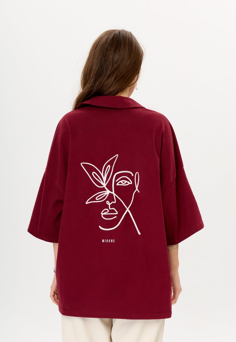 Bordeaux oversized shirt met een geknolde kraag. Heeft witte geborduurde lijntekening van een gezicht en een blad op de achterkant. Tekst "MIRONS" onder het ontwerp.