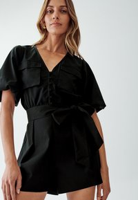 Jumpsuit negro con escote en V, manga corta, botones frontales, dos bolsillos en el pecho y un cinturón de tela en la cintura. Textura suave.