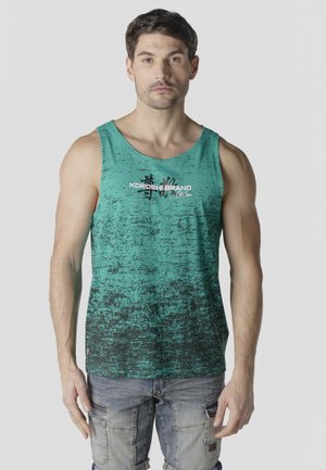 Hombre con cabello corto castaño y barba ligera, lleva una camiseta sin mangas moteada en verde azulado y negro, y pantalones cortos cargo desteñidos, de pie contra un fondo gris.