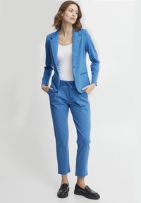 Klart blå skräddarsydd blazer och matchande byxor, tillverkade av mjukt tyg, med en enda knapp, fickor och resår i midjan. Svarta loafers.