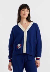 Blauwe cardigan met een crèmekleurige V-hals, knopen aan de voorkant, twee zakken met een ronde patch en gestreepte manchetten in rood, wit en blauw.