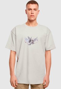 Svetle šedé oversized tričko s centráln�ím motivem bílé ptáka s roztaženými křídly, obklopeného kruhovým fialovým akcentem.