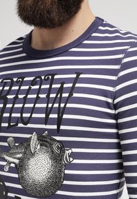 Camisa de manga larga a rayas en púrpura y blanco, con un gráfico negro de un pez y texto en negrita. Fabricada en una tela suave y elástica.