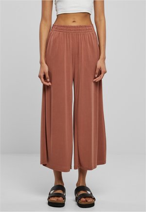 Femme portant un pantalon ample à jambes larges de couleur rouille avec une taille élastique et des sandales noires à enfiler, debout devant un fond gris.