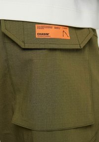 Olivově zelené kalhoty s texturovanou tkaninou; mají velkou, sklopenou zadní kapsu a oranžovou etiketu s nápisem "CHASIN' PERFORMANCE JEANSWEAR."