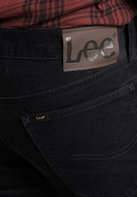 Černé kordové kalhoty mají výraznou koženou nášivku s vyraženým nápisem "Lee" v metalickém písmu. Malá zlatá nášivka "Lee" na kapse.