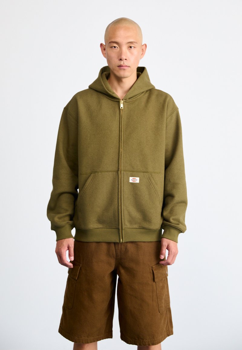Hoodie de fleece verde oliva com zíper completo, grandes bolsos frontais e punhos canelados, acompanhado de calções cargo castanhos com bolsos laterais.