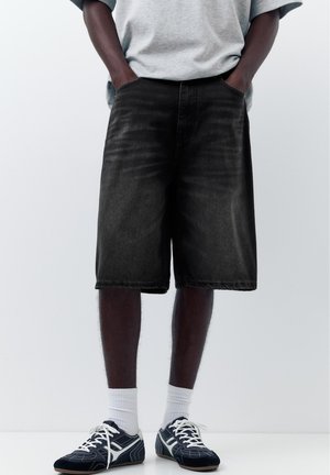 Person trägt graues T-Shirt, schwarze knielange Jeans-Shorts, weiße Sportsocken und schwarz-weiße Sportschuhe, Hände in den Taschen.