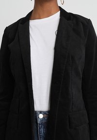 Kobieta ubrana w czarną marynarkę na białej koszulce i niebieskich dżinsach, z złotymi kolczykami w kształcie koła, na neutralnym tle.