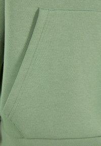 Tissu de sweat-shirt vert avec une texture lisse. Comprend une poche kangourou à l'avant et des poignets côtelés au bas et aux extrémités des manches.