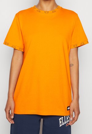 Person iført en knaldorange t-shirt med korte ærmer med små sorte halvcirkelsmønstre på kraven og ærmerne, kombineret med marineblå shorts.