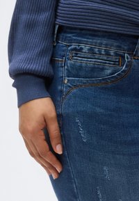 Sötétkék mosású farmernadrág, látható varrással, karcsúsított szabással és finom kopott hatású részletekkel az elülső zsebrészen.