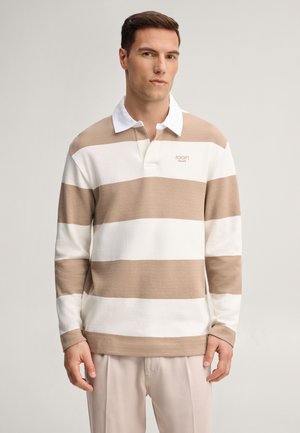 Homme portant un polo de rugby à manches longues à rayures beige et blanches avec un col blanc et un pantalon beige, debout devant un fond uni.