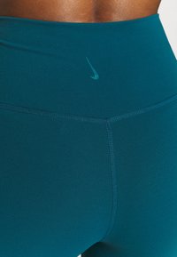 Teal högmidjade träningsleggings tillverkade av ett mjukt tyg, med platta sömmar och en liten Nike-logotyp på framsidan.