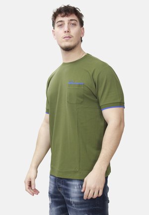 Giovane uomo che indossa una camicia verde oliva a maniche corte con bordino blu e taschino sul petto, abbinata a jeans blu strappati, che posa su sfondo bianco.