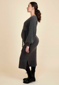 Graues Strickkleid mit langen Puffärmel, einem Bindeband in der Taille und einem seitlichen Schlitz. Kombiniert mit schwarzen Stiefeletten und Strumpfhosen.