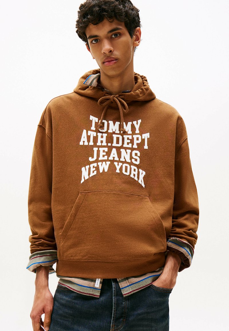 Sweat à capuche marron avec le texte blanc "TOMMY ATH.DPT JEANS NEW YORK," doté d'une poche avant et superposé avec une chemise rayée multicolore en dessous.