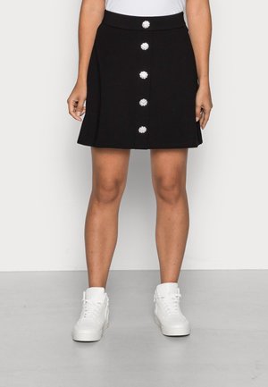 Mujer con falda negra con cinco grandes botones decorativos en la parte delantera y zapatillas blancas altas de lona, de pie sobre un suelo gris.
